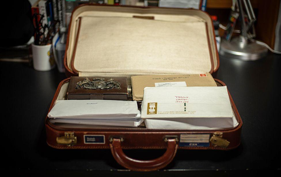 Uma mala de couro marrom aberta repousa sobre uma superf&iacute;cie escura, contendo uma caixa de metal decorado, um pacote envolto em papel pardo e v&aacute;rios envelopes brancos. Um dos envelopes est&aacute; marcado com a inscri&ccedil;&atilde;o 