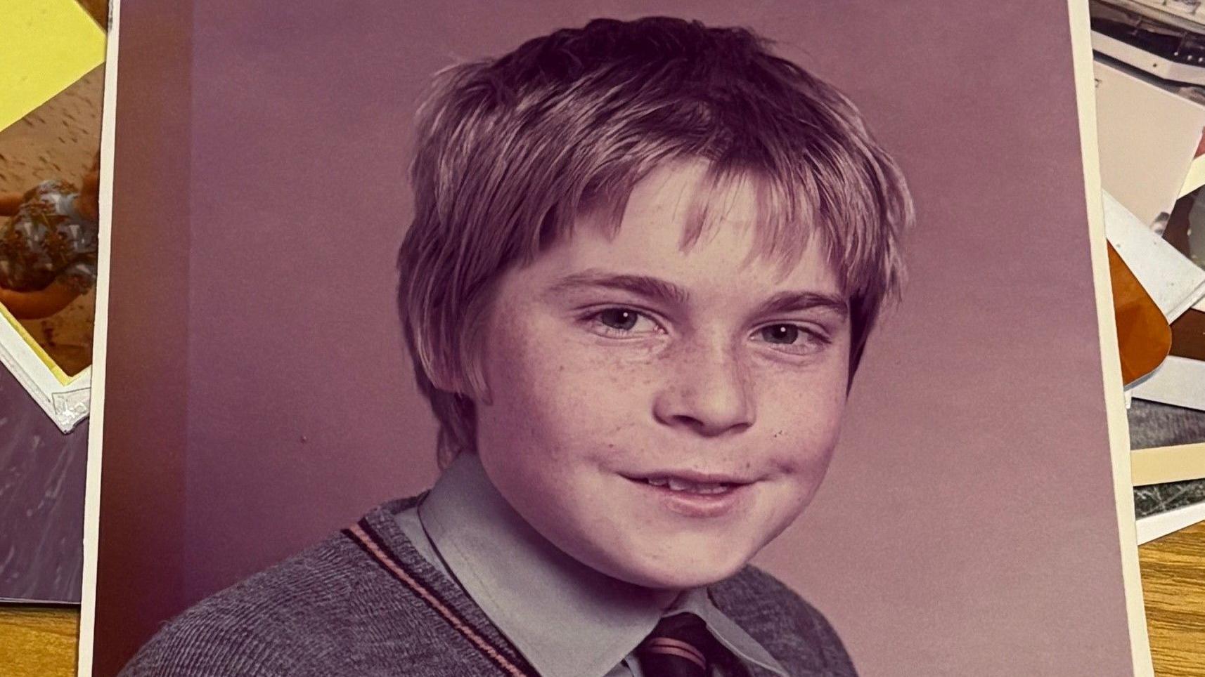 Uma antiga fotografia escolar mostra um menino com sardas. Ele veste uma camisa cinza e uma gravata preta e dourada sob um su&eacute;ter grande de gola em V