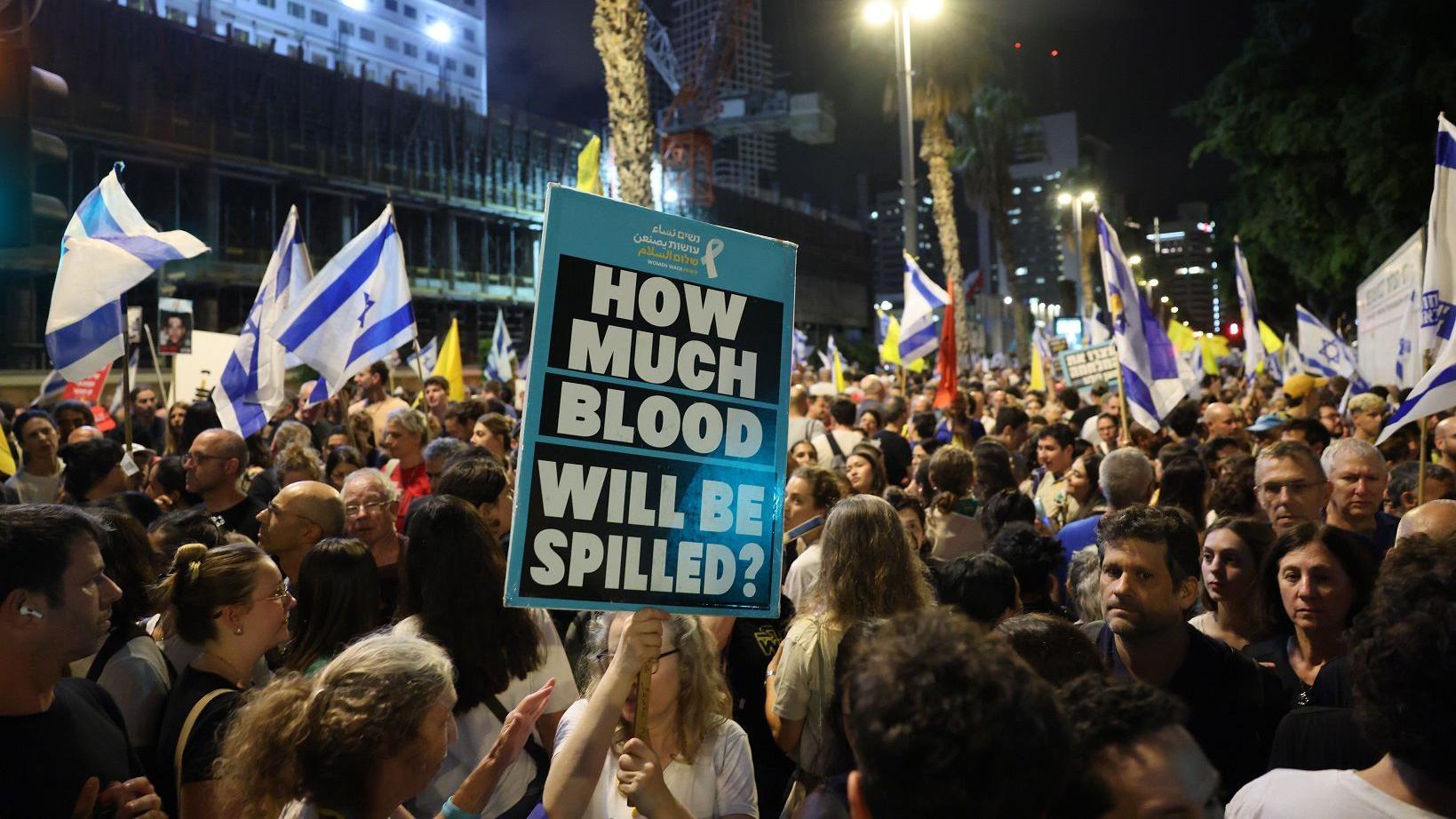 Pessoas na rua durante manifestação, com bandeiras de Israel e um cartas perguntando, em inglês, 