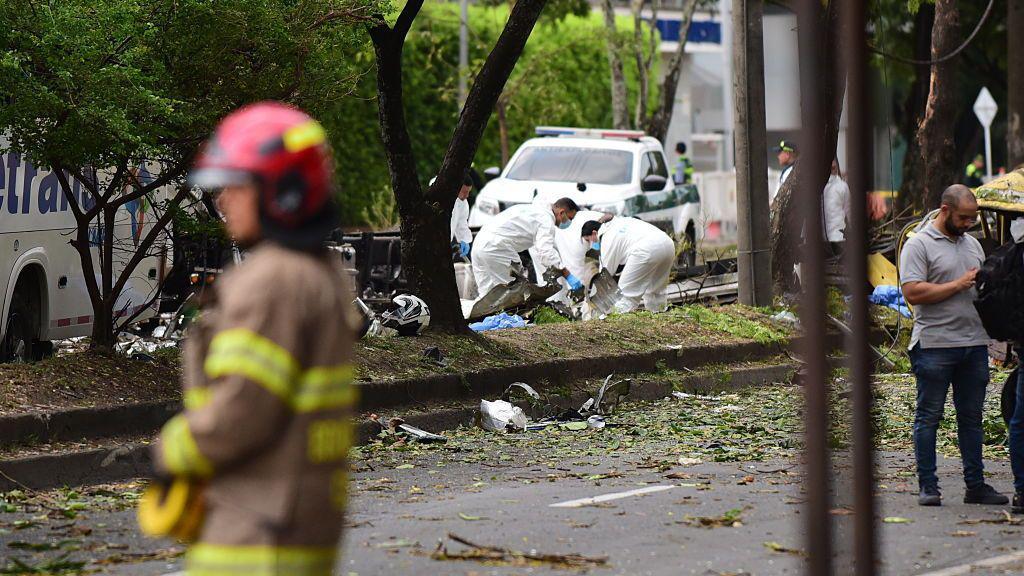 Las autoridades revisan la escena en donde explotó un camión bomba en Cali, colombia. Al fondo se ven varios funcionarios forenses revisando el suelo y un bombero en primer plano. 