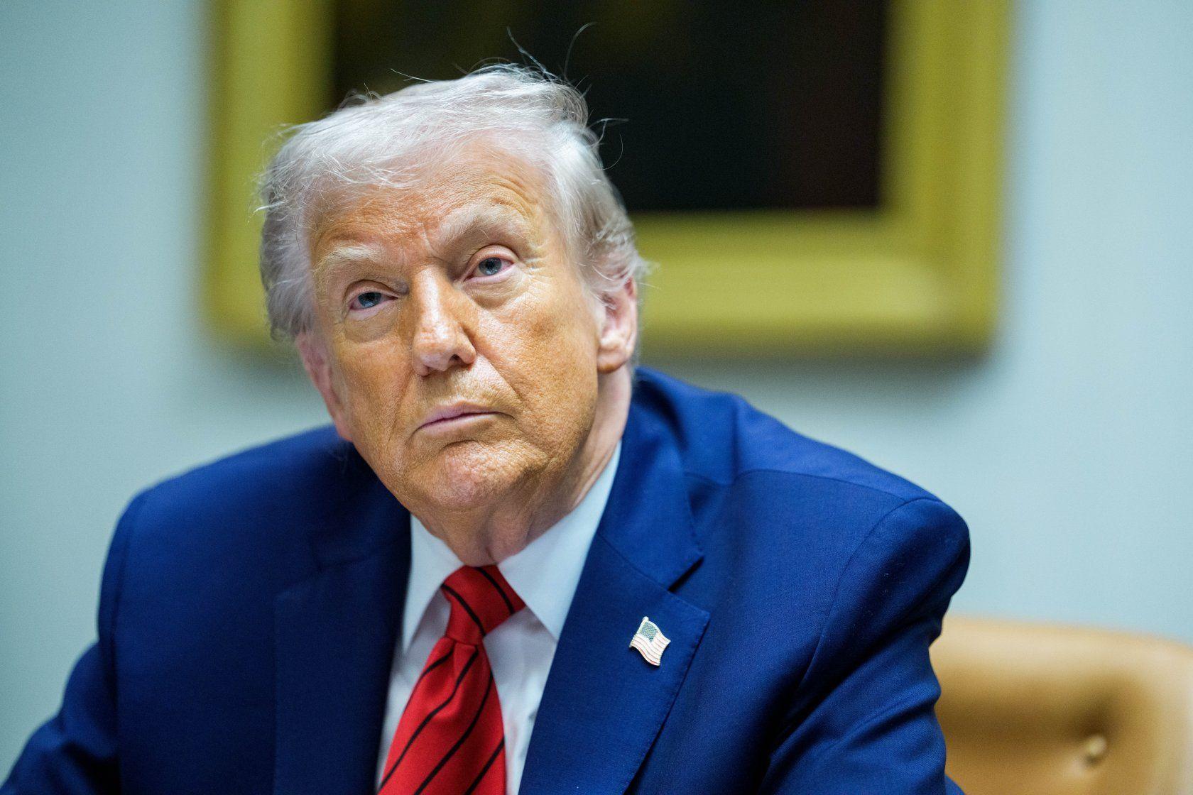 美國總統特朗普，坐姿，身穿藍色西裝、襯衫、紅色條紋領帶，並佩戴美國國旗徽章於西裝翻領上