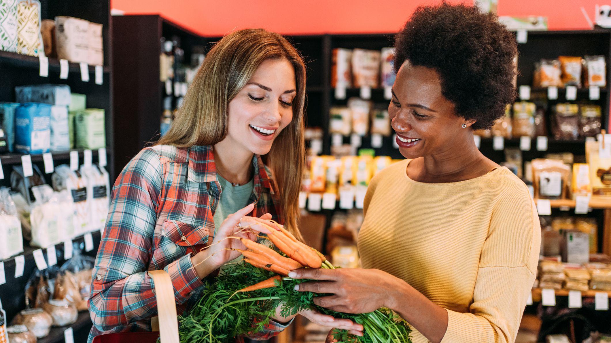 Duas mulheres conversam e escolhem cenouras no supermercado