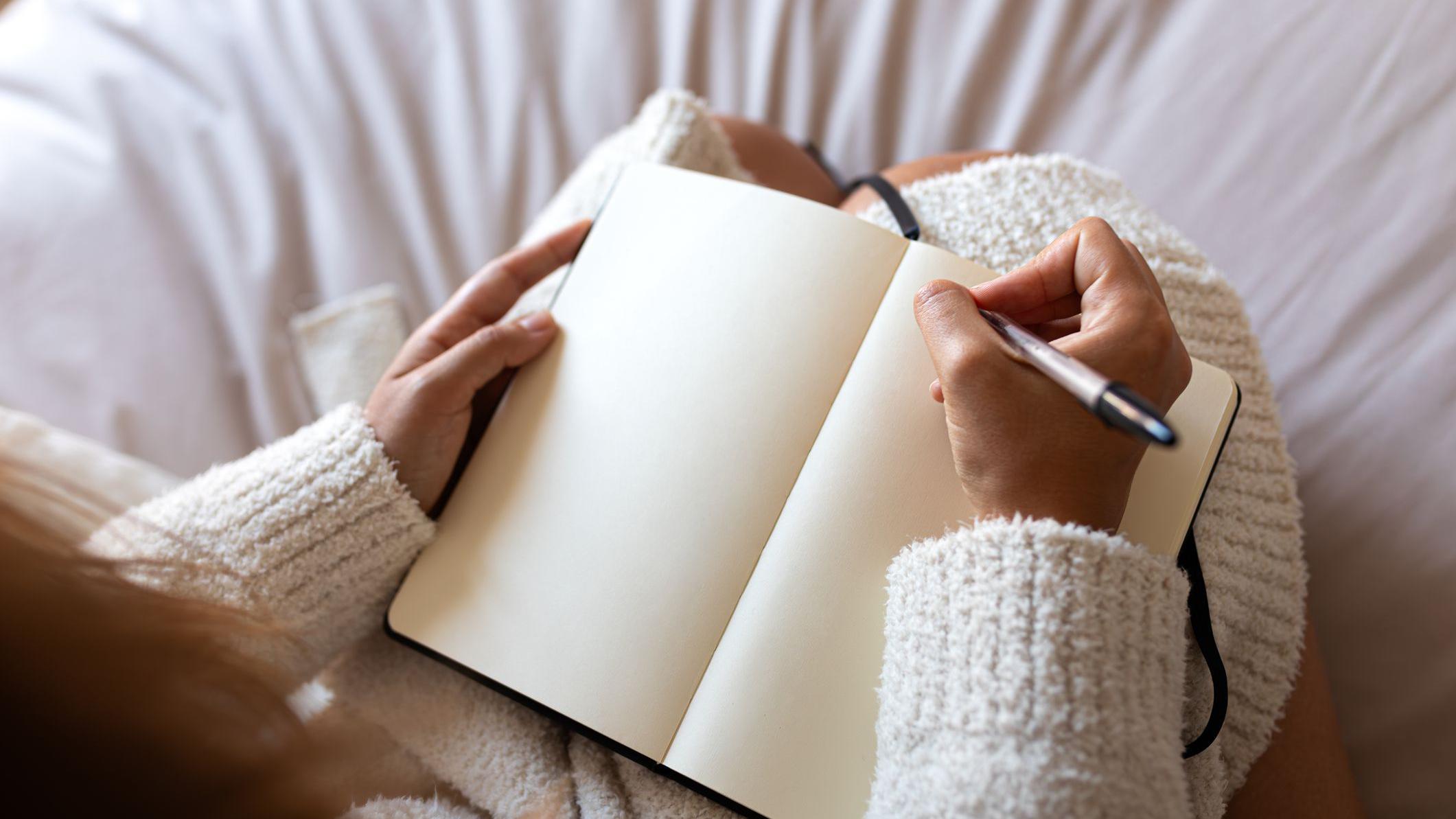 Vista em &acirc;ngulo de cima de uma pessoa, vestindo um roup&atilde;o, sentada na cama escrevendo em um di&aacute;rio em um quarto aconchegante.