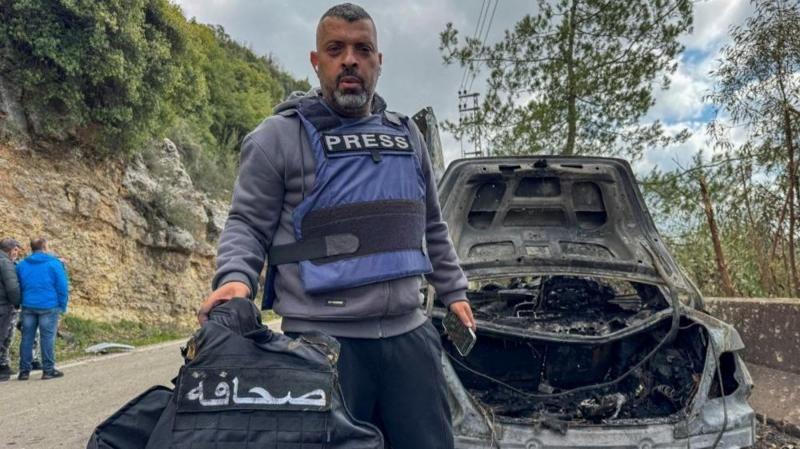Un hombre con un chaleco de protección con un rótulo que dice "prensa" muestra a la cámara otro chaleco junto a un auto carbonizado. 
