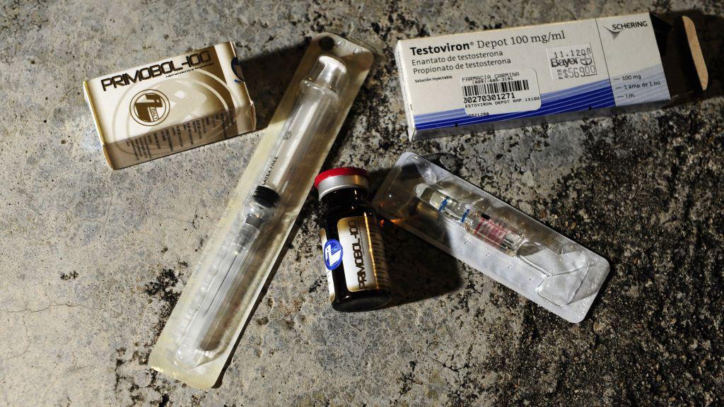 A imagem mostra diferentes itens associados ao uso de esteroides anabolizantes e testosterona dispostos sobre uma superfície de concreto. Entre eles, há duas caixas de medicamentos — uma identificada como Primobol-100 e outra como Testoviron Depot 100 mg/ml —, um frasco de vidro com tampa vermelha e duas seringas embaladas individualmente em plástico estéril. A composição destaca os produtos e os materiais utilizados para aplicação injetável dos hormônios.