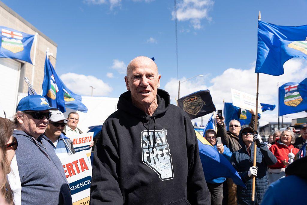 Mitch Sylvestre, director de Stay Free Alberta, un hombre de mediana edad, con poco pelo, viste una sudadera negra que lee "Queda libre" sobre el mapa de la provincia de Alberta. Está rodeado de simpatizantes que ondean banderas de Alberta