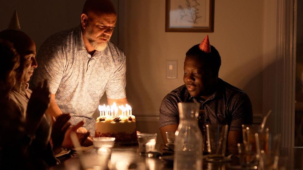 Marven observa con una sonrisa cómo un hombre lleva un pastel de cumpleaños con velas encendidas a la mesa donde él y otras personas están sentados. Las luces son tenues y Marven lleva un sombrero de fiesta.