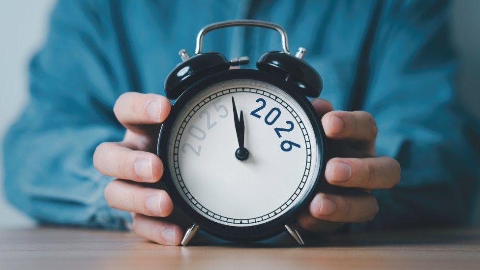 Joven sosteniendo un reloj despertador en el que las agujas están en transición entre 2025 y 2026