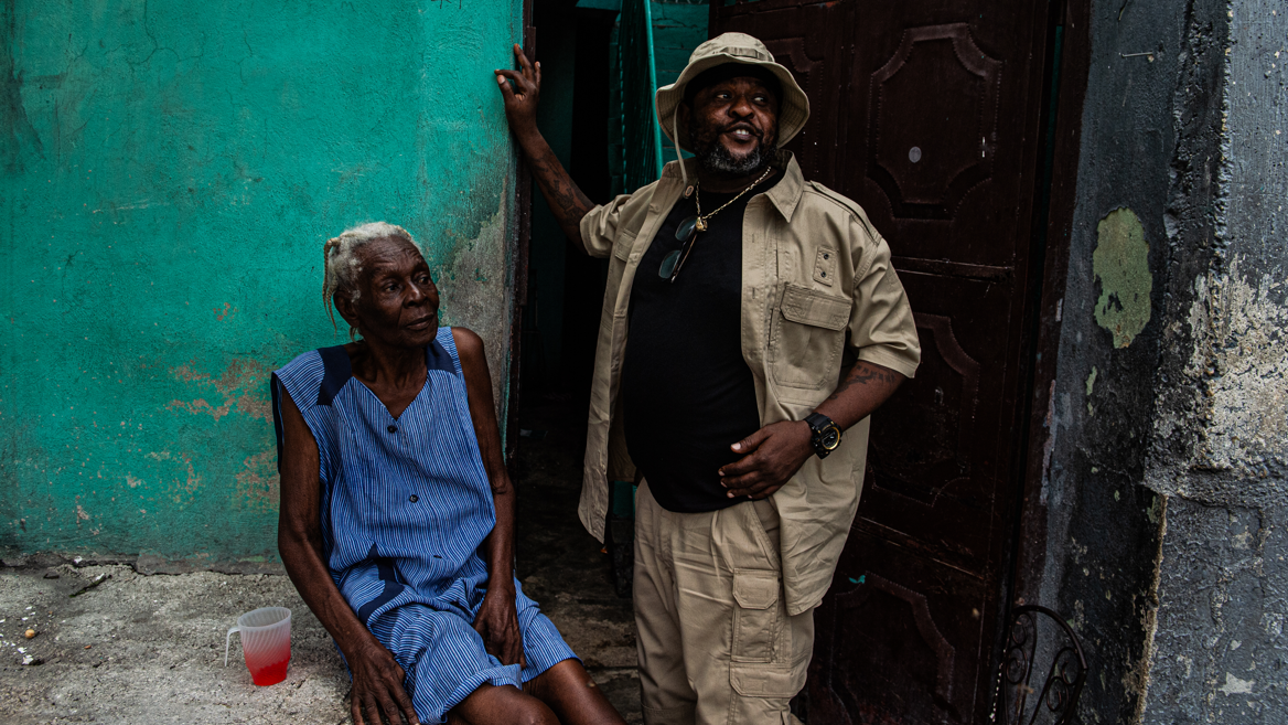 O l&iacute;der da Viv Ansanm Jimmy Ch&eacute;rizier, conhecido como Barbecue, de camisa, cal&ccedil;a e chap&eacute;u bege, ao lado de uma anci&atilde; vestida de azul, em um bairro de Porto Pr&iacute;ncipe, no Haiti, em 6 de julho de 2026