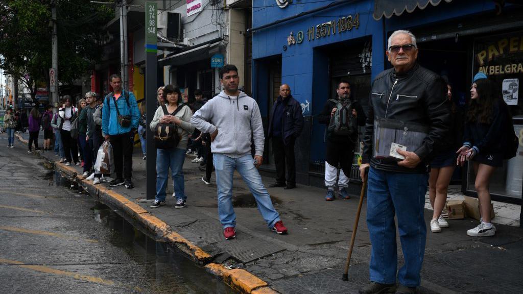 Um grupo de pessoas espera um ônibus parado na calçada de uma avenida em Buenos Aires.