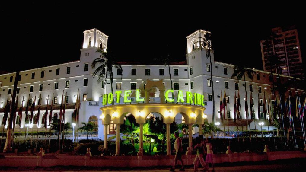 Una vista nocturna del Hotel Caribe de Cartagena, de arquitectura al estilo colonial, rodeado de palmeras y banderas de muchas naciones, iluminado con luces blancas. El nombre del hotel resalta en grandes letras de luz verde.