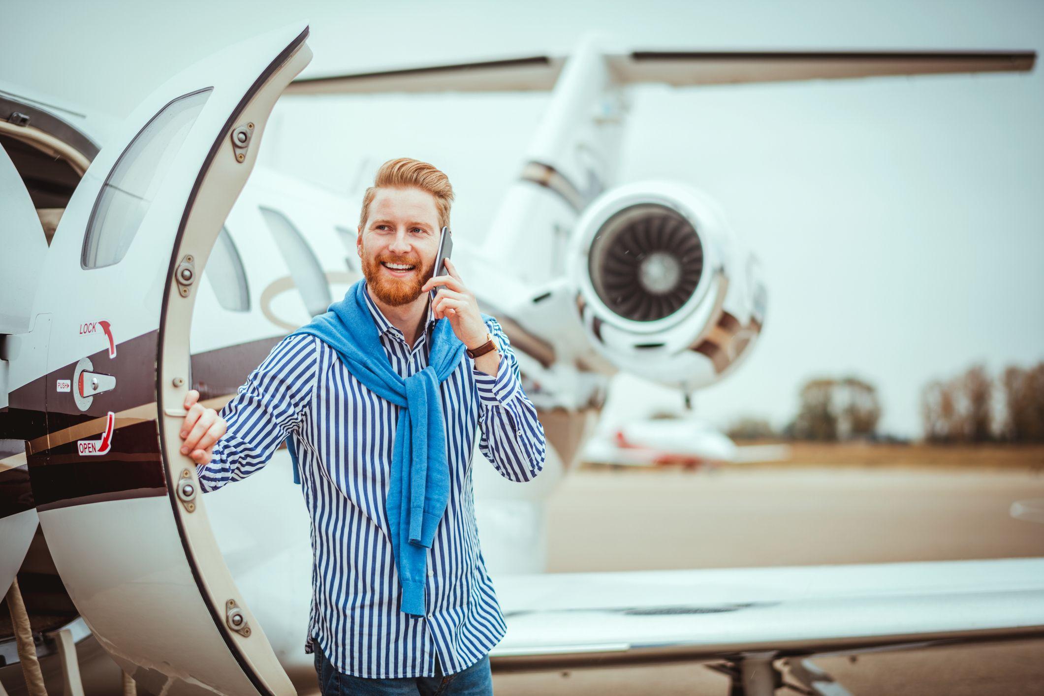 Homem ao celular ao lado de um jato particular estacionado numa pista de pouso