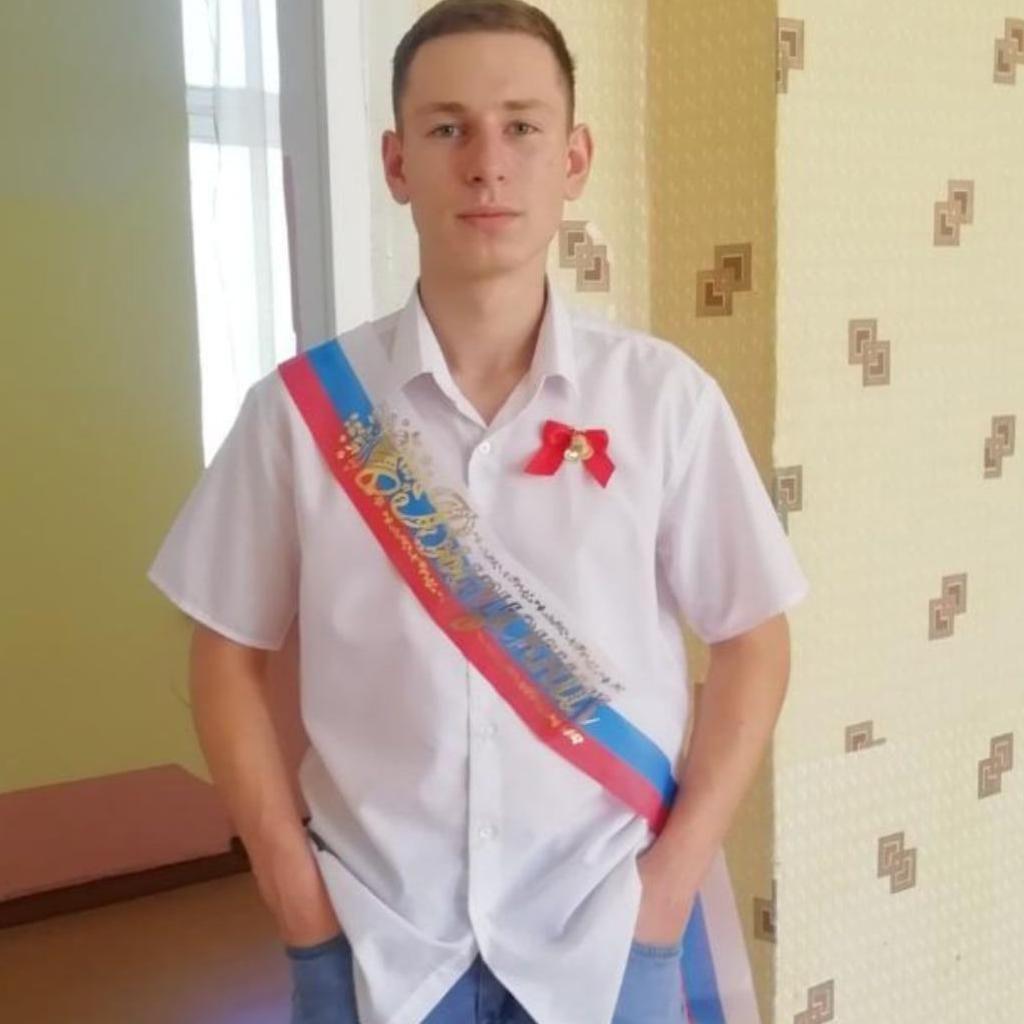 Aleksandr Petlinsky com uma camisa branca, uma faixa na transversal do corpo e um laço vermelho no peito. 