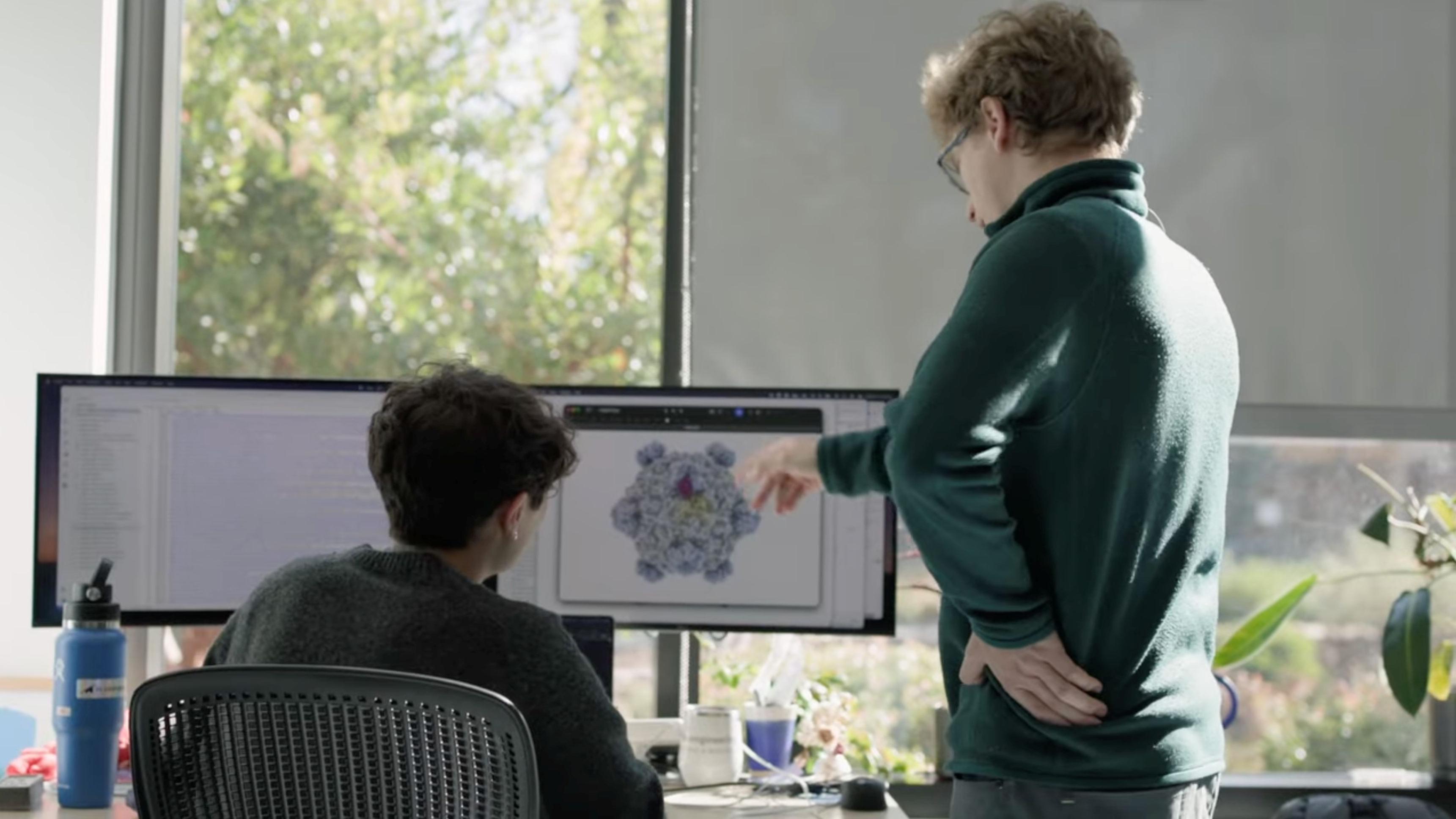 Esta imagen muestra a dos personas trabajando en un escritorio en una oficina o laboratorio luminoso, con grandes ventanales y luz natural. Una persona está sentada frente a una estación de trabajo con dos monitores, mientras que la otra se encuentra cerca, señalando una estructura molecular o proteica detallada que se muestra en una de las pantallas. Los monitores muestran software de visualización científica: una pantalla presenta un complejo modelo biológico tridimensional y la otra contiene texto o datos. Se pueden observar diversos objetos alrededor del escritorio, como una botella de agua, una taza, periféricos de computadora y plantas en macetas. 