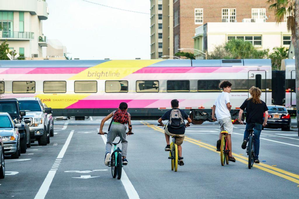 Cuatro niños montan en bicicleta por una calle por la que cruza un tren Brightline. 