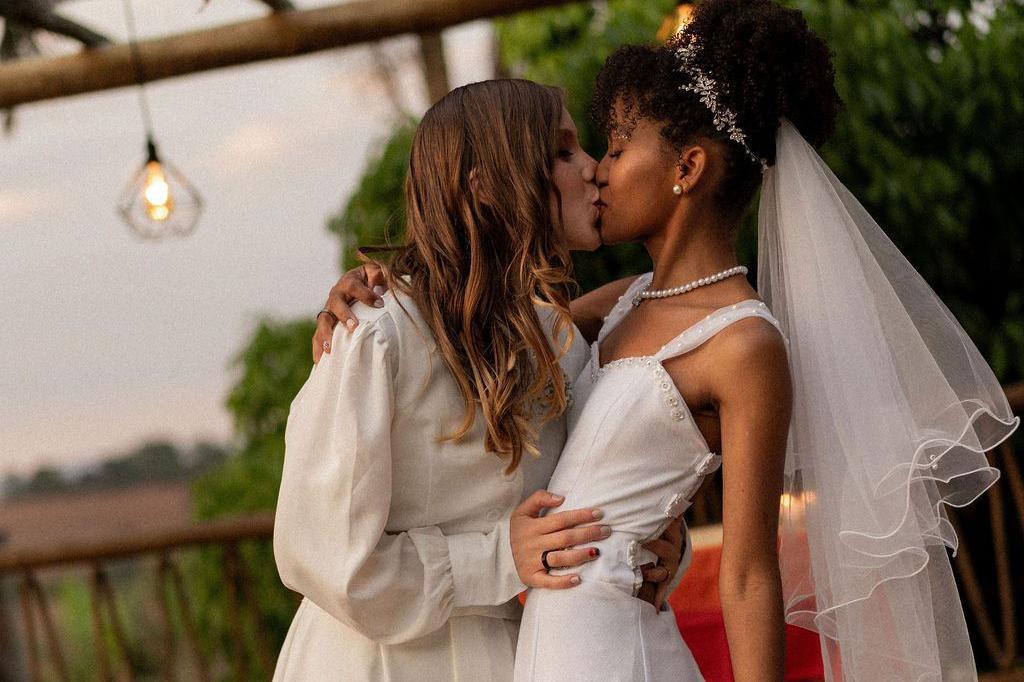 Fotografia colorida mostra duas mulheres vestidas de branco se beijando durante cerim&ocirc;nia de casamento
