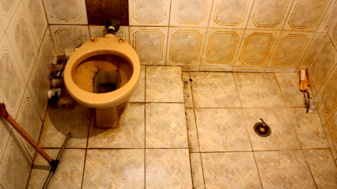 Banheiro com algumas partes da parede sem azulejo, enquanto outras t&ecirc;m azulejo manchado; privada sem tampa e suja