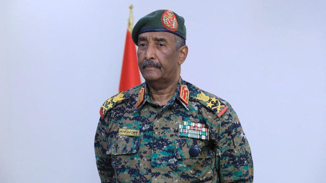 عبد الفتاح البرهان يرتدي زياً عسكرياً مموهاً مع قبعة خضراء، يقف أمام خلفية بيضاء مع جزء من العلم السوداني ظاهر في الخلفية.