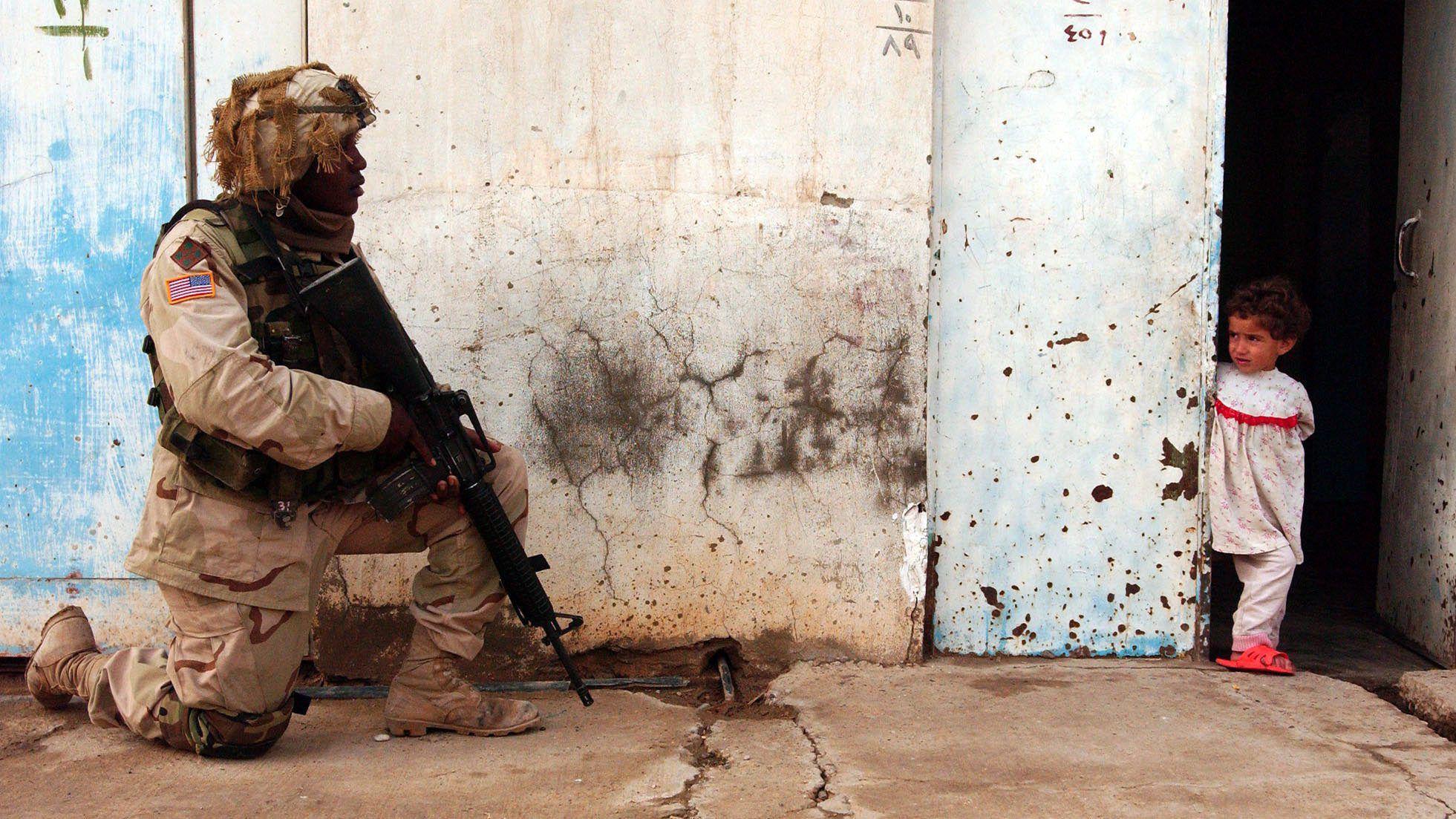 Američki vojnik čuva ulicu tokom patrole, kleči na betonu sa puškom uperenom nadole. Nosi kamuflažnu uniformu. Devojčica stoji iza ugla zgrade i gleda ga.