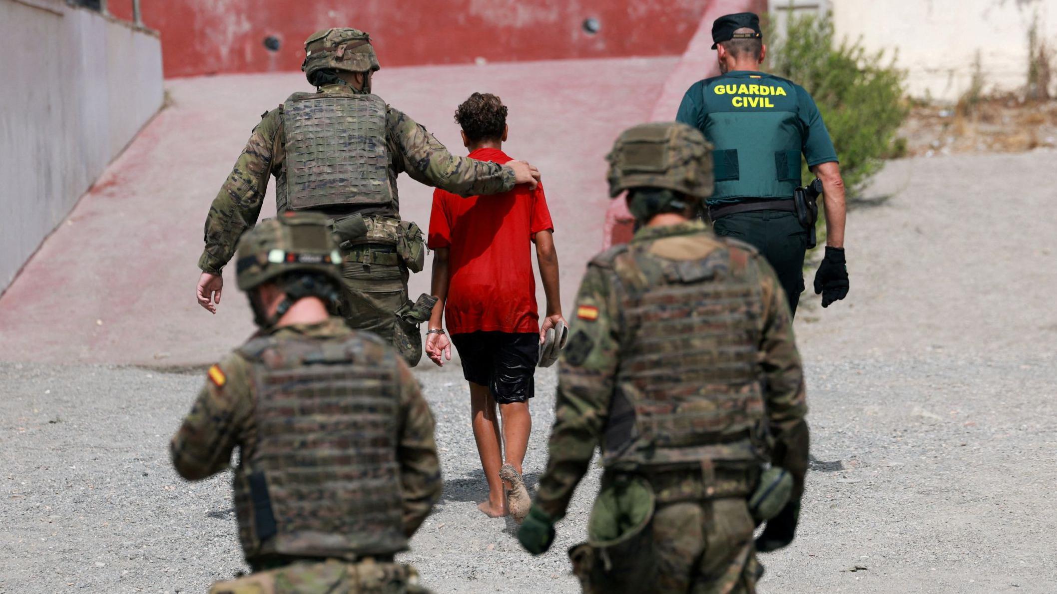 Tres soldados y un agente de la Guardia Civil acompañan a un menor con una camiseta roja y pantalones cortos. Están todos de espaldas. Ceuta, 1 de agosto de 2026