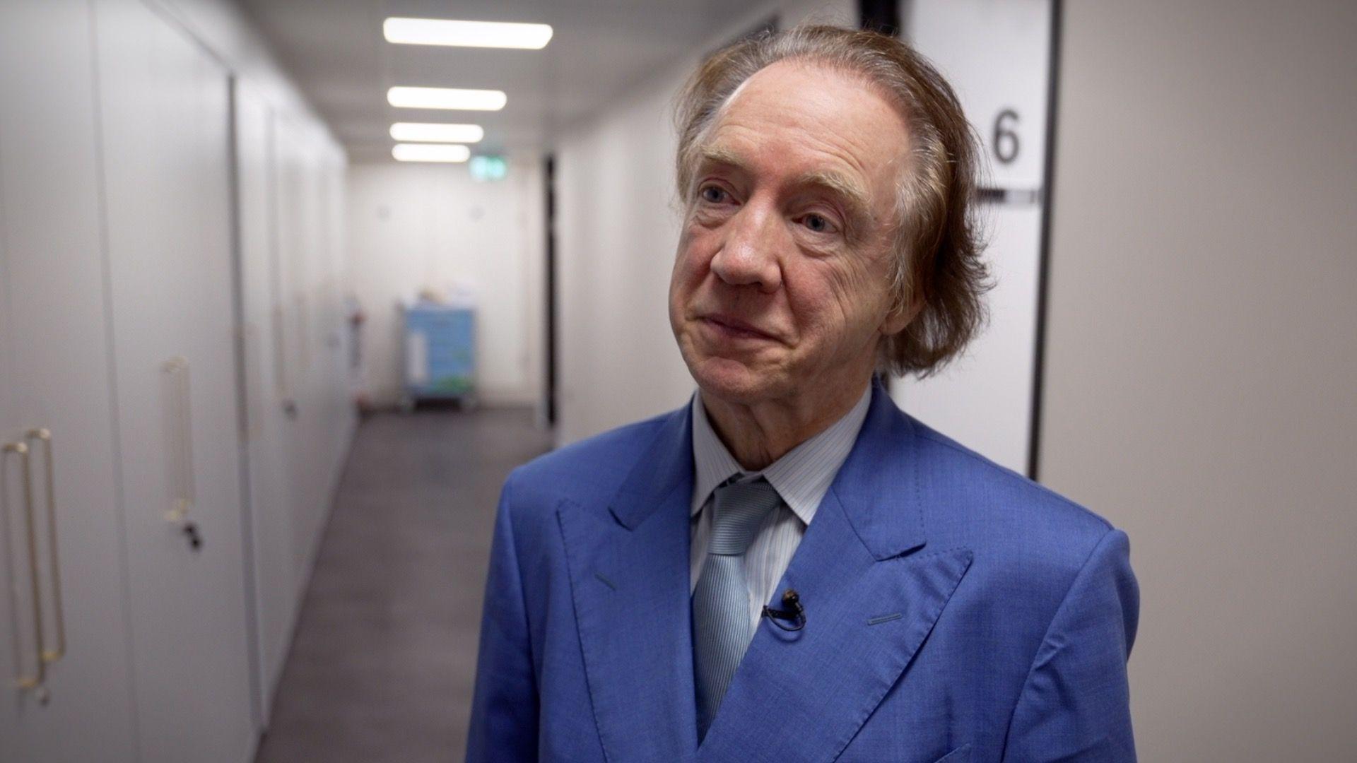 一名年長男子身穿藍色西裝，搭配襯衫和淡藍色領帶。他的棕色頭髮向後梳理，視線投向右側。