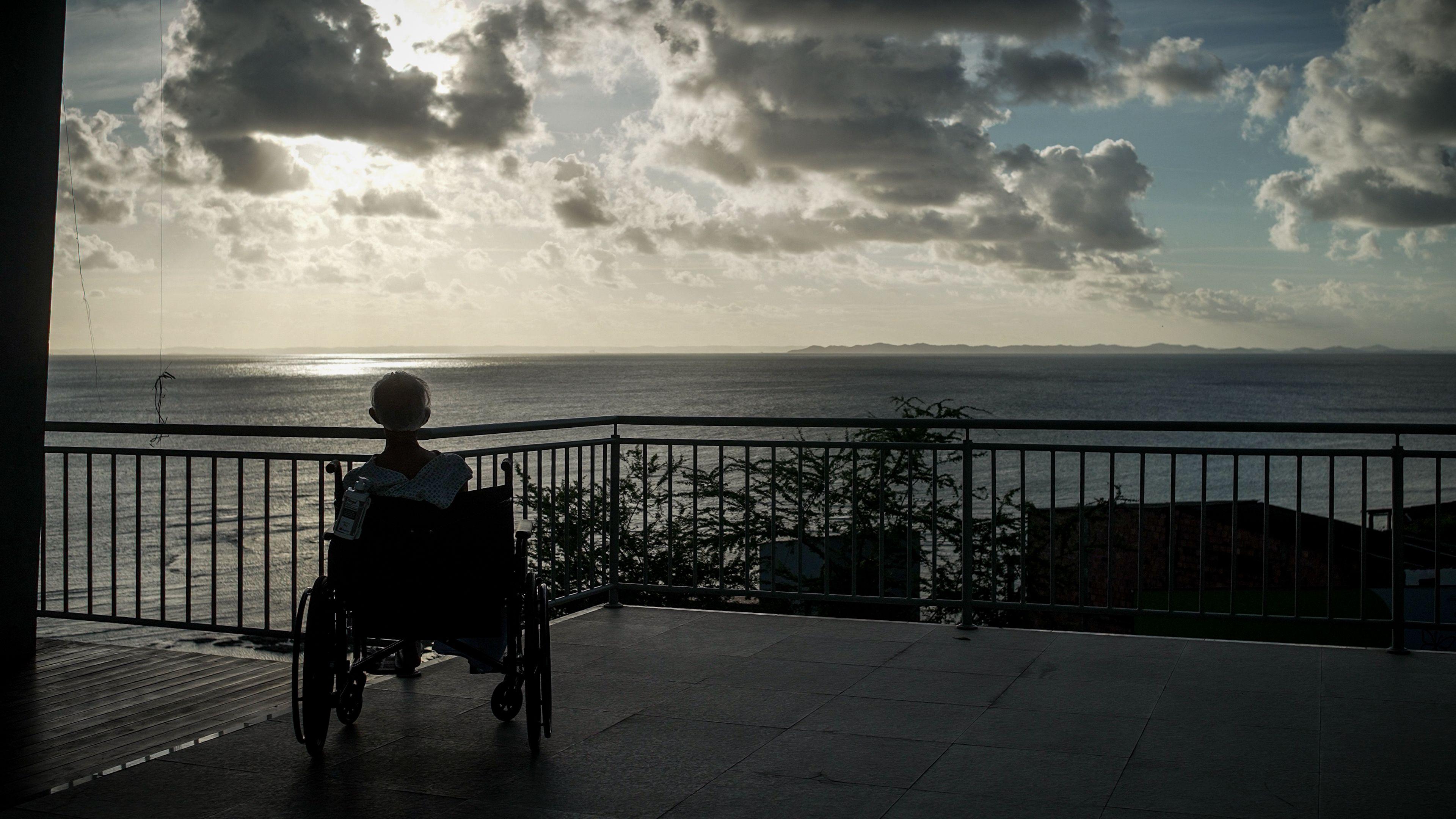 Foto de um senhor em uma cadeira de rodas de costas para a c&acirc;mera enquanto contempla o p&ocirc;r do sol em um p&iacute;er sobre o mar.