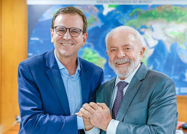 Paes, de blusa e blazer azuis, posa ao lado de Lula, que usa terno cinza e gravata estampada roxa. Ambos sorriem e d&atilde;o as m&atilde;os em um gesto de alian&ccedil;a