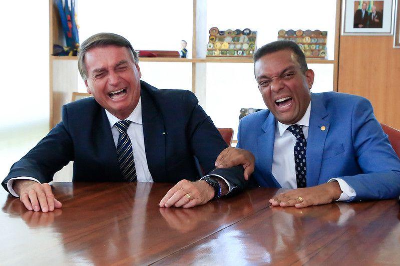 De terno preto e gravata listrada, Bolsonaro, est&aacute; sentado ao lado de Otoni de Paula, que usa terno azul, gravata estampada e segura o bra&ccedil;o do ent&atilde;o presidente. Ambos gargalham. 