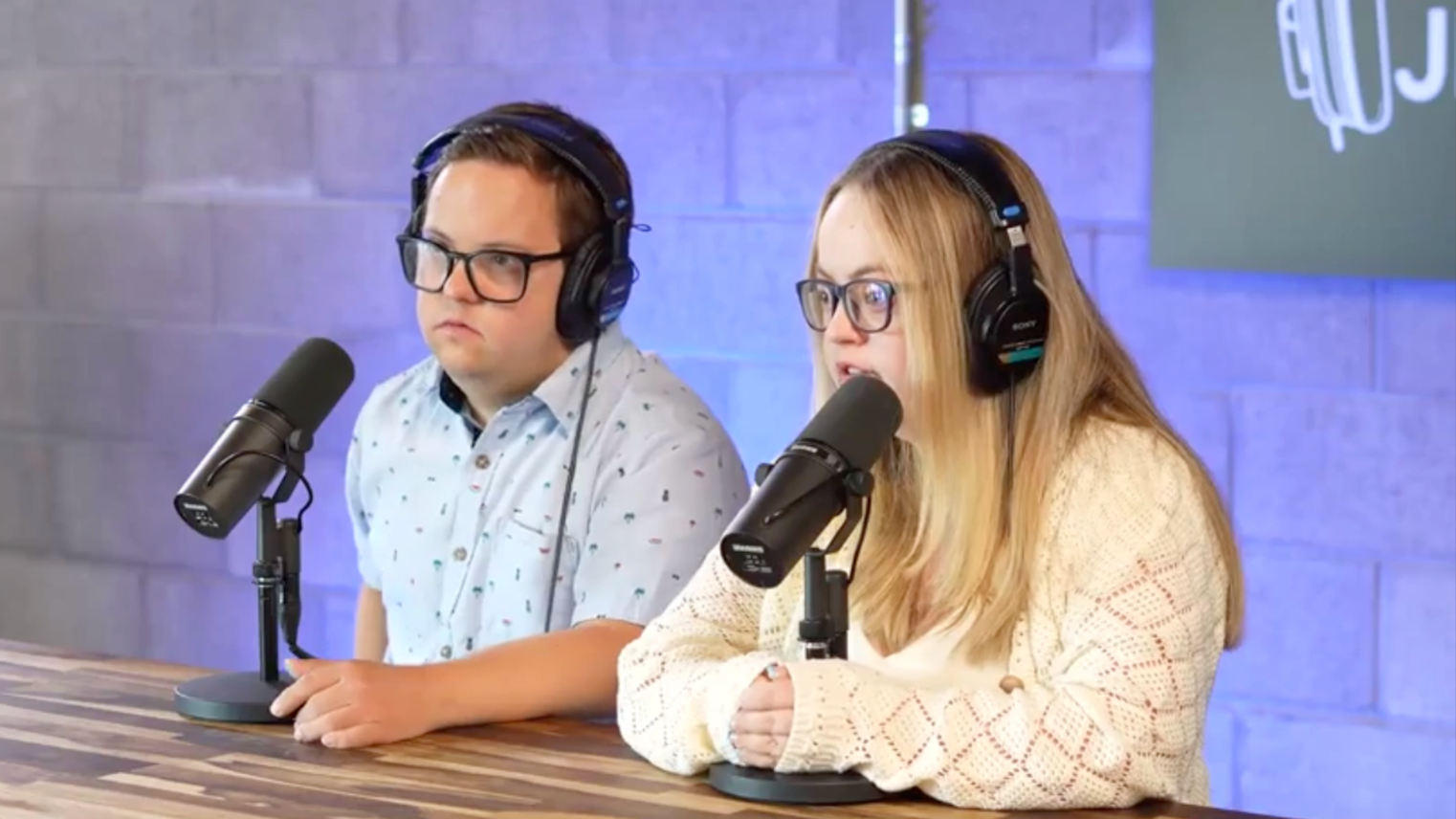 O jovem casal Audrey e Jeremy, sentados atrás de uma mesa de madeira, usando fones de ouvido e falando ao microfone. A iluminação é azul e a sala parece profissional. 