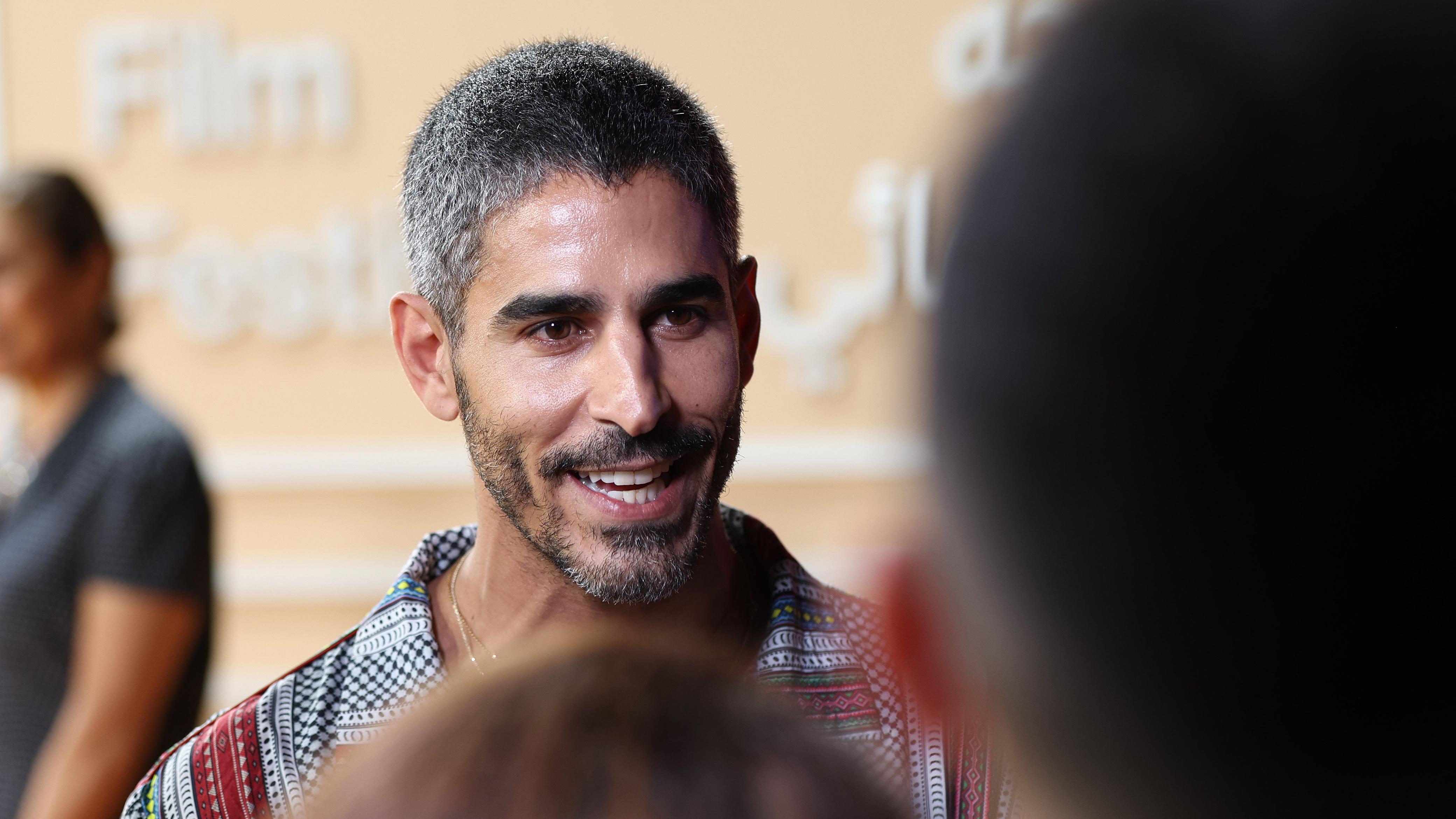 Ahmed Shihab‑Eldin en la noche de apertura del Festival de Cine de Doha 2025. Sonríe mientras conversa con personas cuyos rostros aparecen desenfocados en primer plano. Una mujer se encuentra de pie a lo lejos.