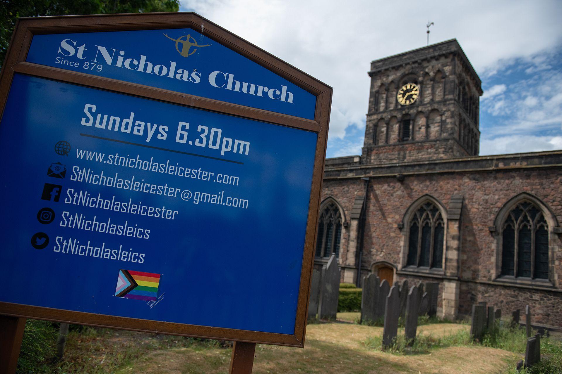 Placa azul com informa&ccedil;&otilde;es sobre missas e detalhes de contato, com a Igreja de S&atilde;o Nicolau de Leicester ao fundo