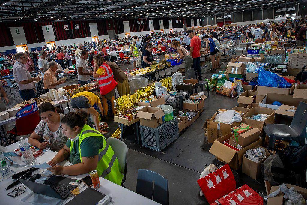 Un enorme centro de refugio para las personas evacuadas. Se ven muchísimas cajas con insumos básicos, como mantas protectoras, comida y demás. Hay muchas personas voluntarias que visten chalecos reflectantes organizando el lugar y entregando insumos. También se ve mucha gente haciendo cola para recoger cosas o esperando al fondo. 
