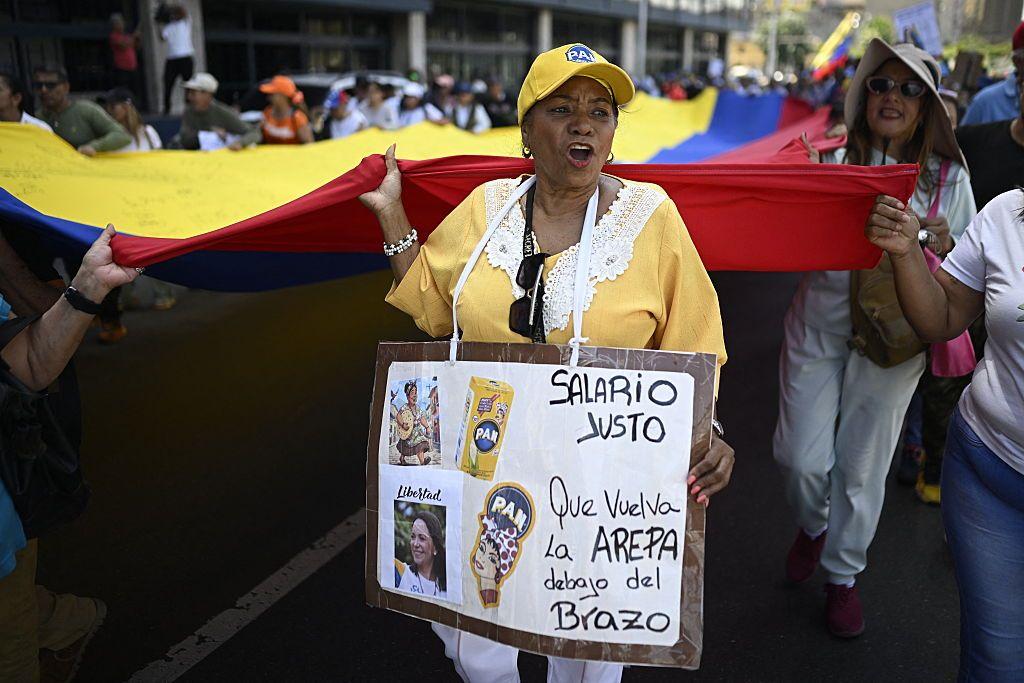 Una mujer con una blusa amarilla participa en una manifestación mientras carga una pancarta colgada del cuello que dice "Salario justo".