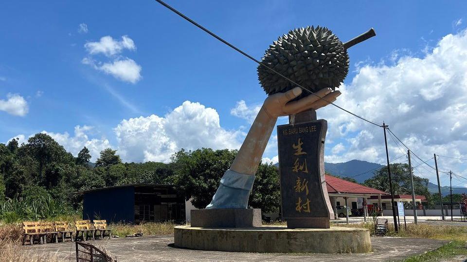 一座巨大的手捧著榴槤的雕像，背景是晴朗的藍天。