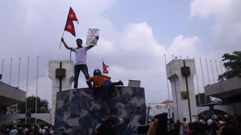Un joven se encuentra en una plataforma con paredes blancas y astas de bandera detrás, el 8 de septiembre de 2025. Sostiene una bandera y un cartel, mientras otra persona sube a la plataforma. La multitud llena el suelo bajo ellos.