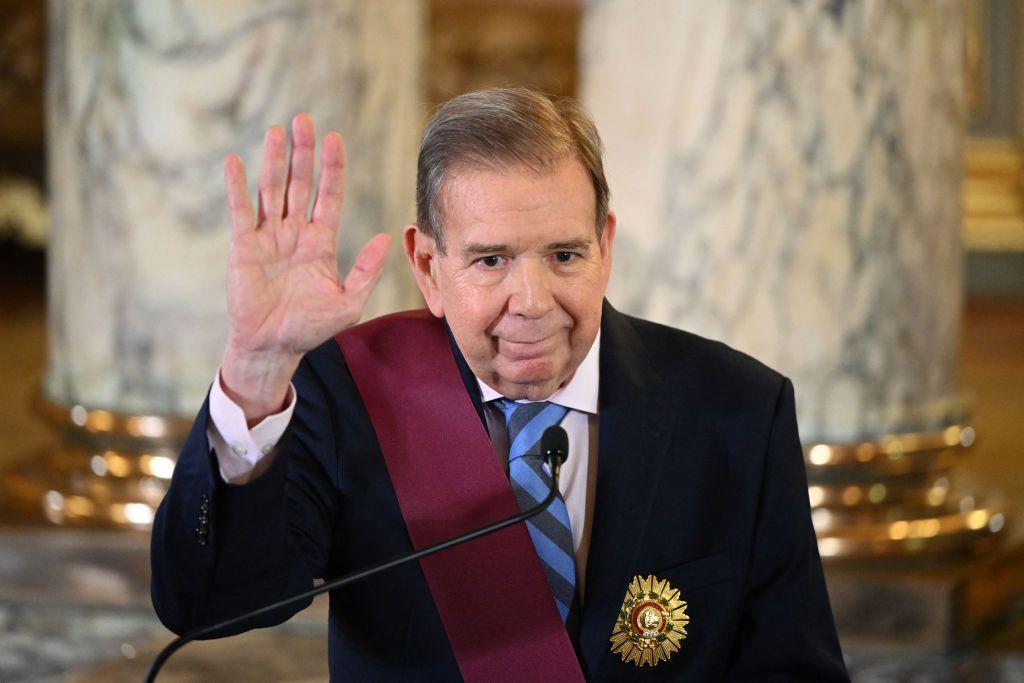 Edmundo González viste una chaqueta azul oscuro, una camisa clara y una corbata de franjas azules y celestes. Tiene la mano derecha en alto como quien está tomando un juramento.