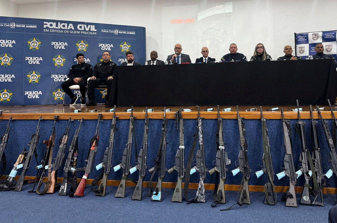 Una serie de rifles capturados por la policía son exhibidos en una conferencia de prensa de la policía civil, mientras los funcionarios hablan detrás.