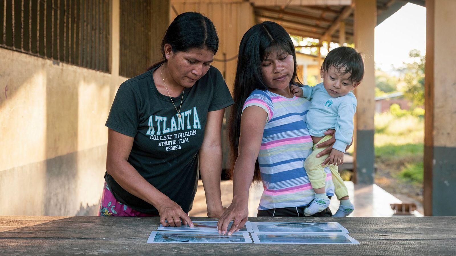 Dos mujeres indigenas en Ecuador señalan fotos de peces. Una de ellas tiene un bebé en brazos. 