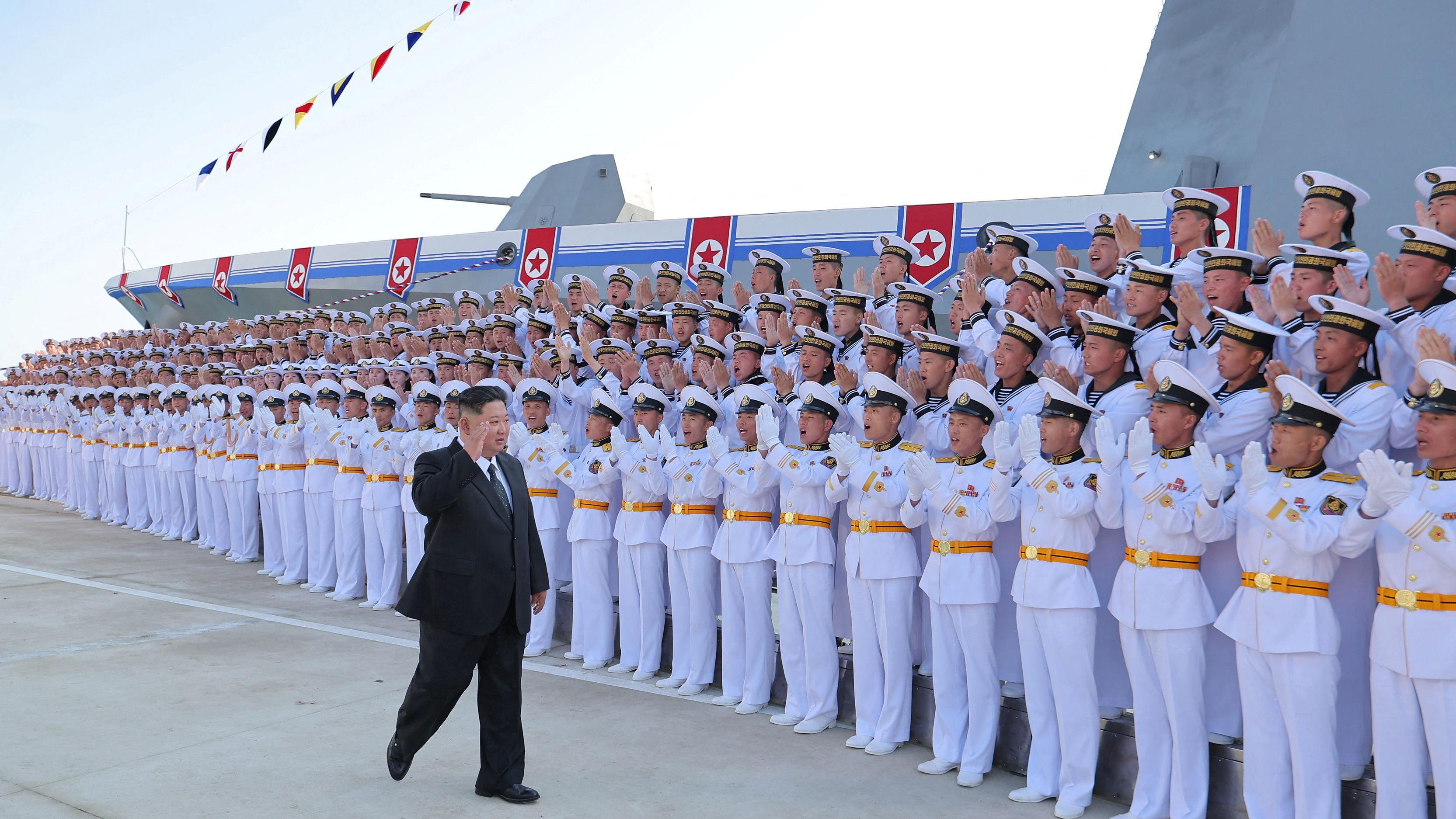 Filas de soldados con uniformes blancos y gorras blancas vitorean mientras Kim Jong Un camina delante de ellos, vestido con un traje negro y levantando la mano. Detrás de ellos hay un enorme barco gris.
