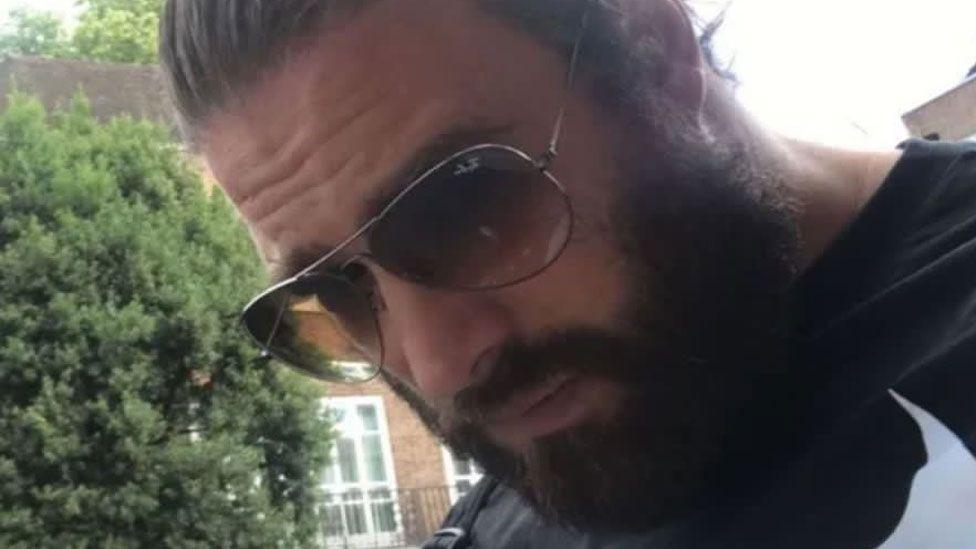 Una foto de Chris Harkins de su perfil de Tinder. Está al aire libre, con árboles y casas al fondo. Lleva el pelo oscuro peinado hacia atrás y barba completa. Usa gafas de sol grandes y tintadas con montura metálica fina. Mira hacia el suelo.