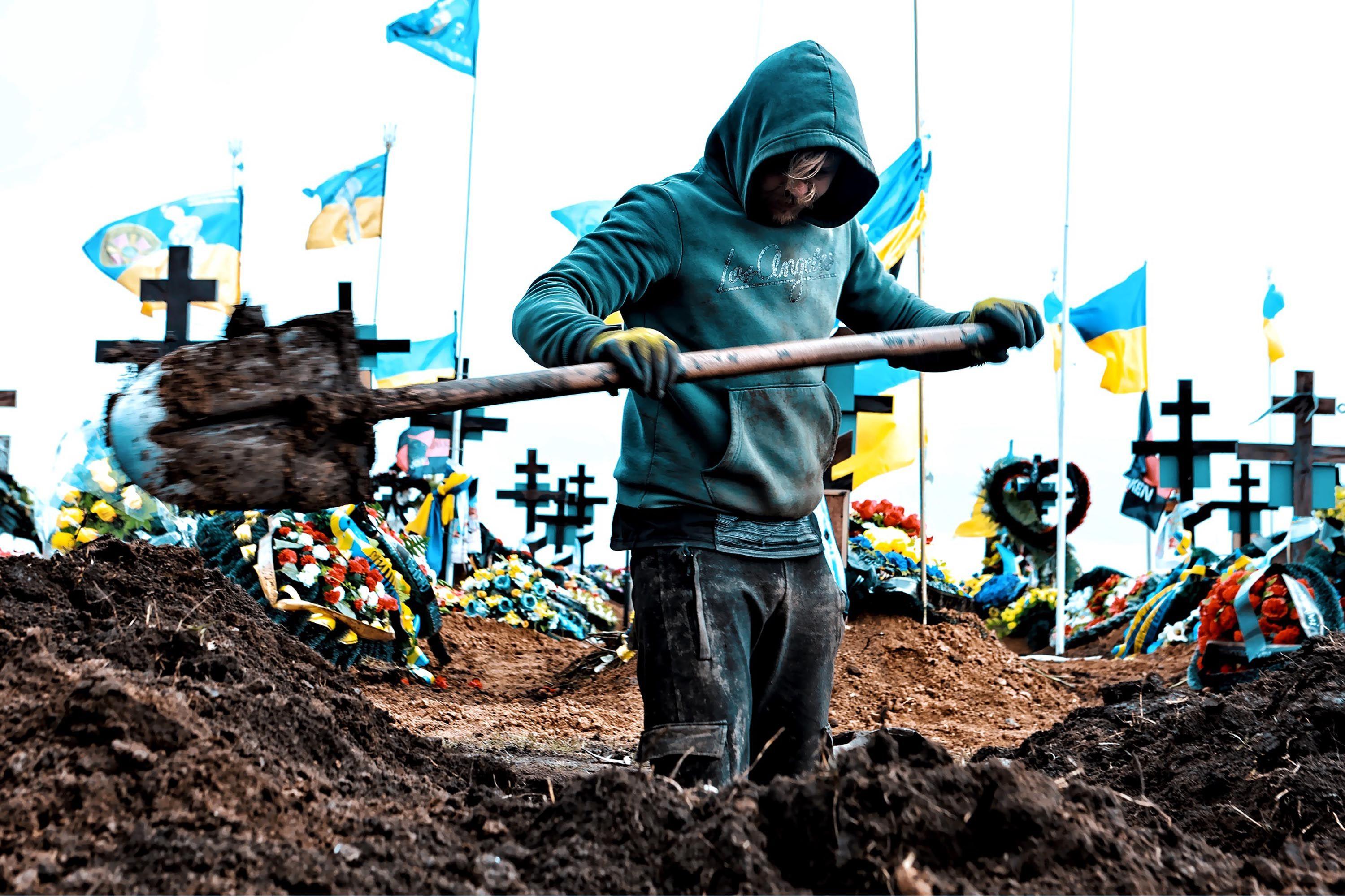 2024年3月16日，掘墓工人在敖德薩的一座墓園挖掘墳墓。他們身後約有十面烏克蘭國旗飄揚在白色旗杆上。前景中的一名掘墓工人身穿青綠色連帽上衣，戴著黃色手套