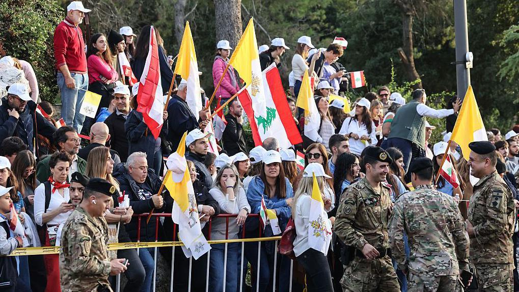 حشود من اللبنانيين من رجال ونساء يقفون على جانب الطريق وخلف حواجز وهم يحملون أعلام الفاتيكان والأعلام اللبنانية للترحيب ببابا الفاتيكان الزائر الباب ليو الرابع عشر.