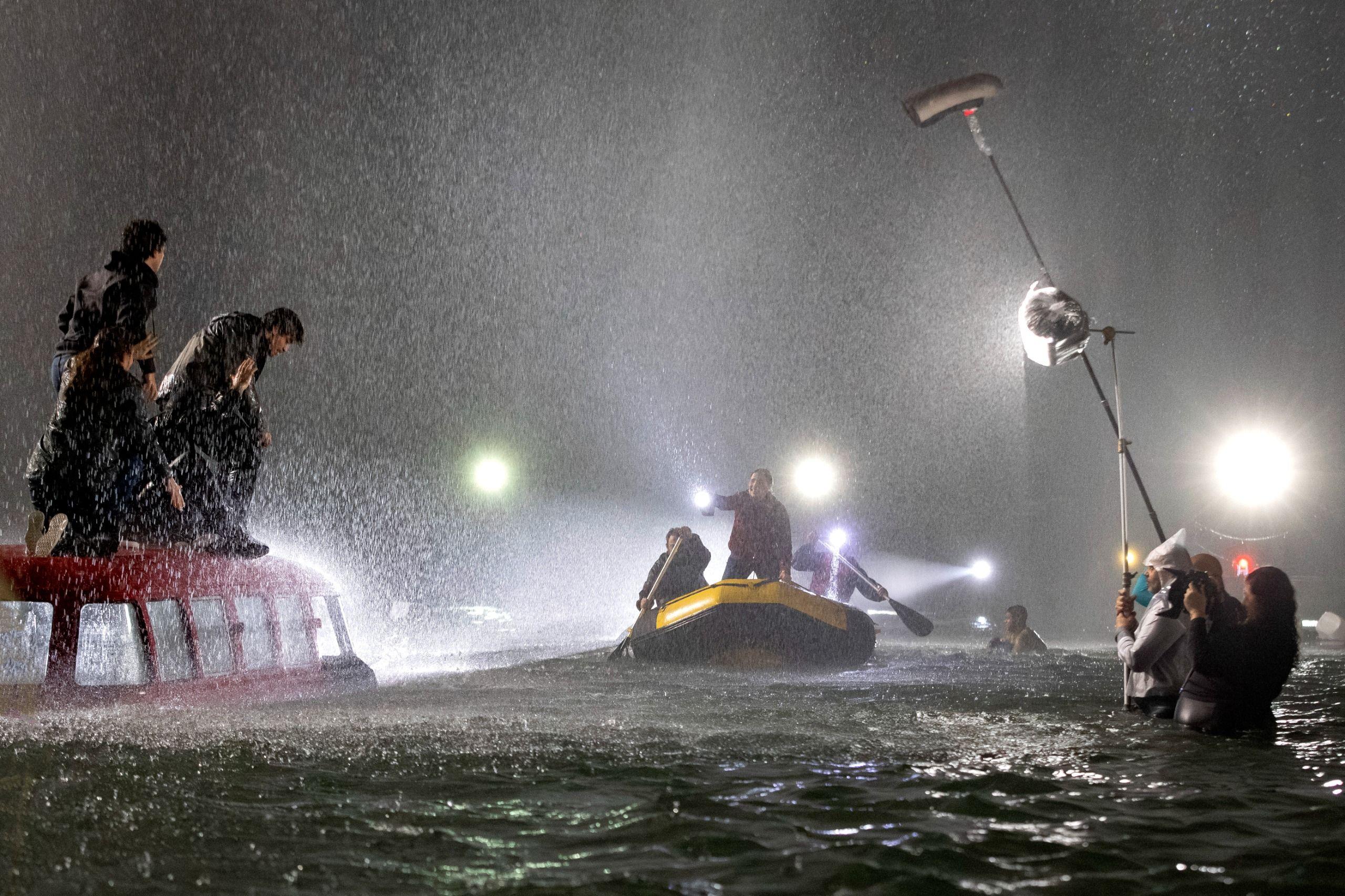 Cena noturna de filmagem na &aacute;gua sob chuva forte: pessoas se equilibram sobre um carro parcialmente submerso enquanto um bote se aproxima, e, ao lado, a equipe opera luz e som dentro da &aacute;gua.