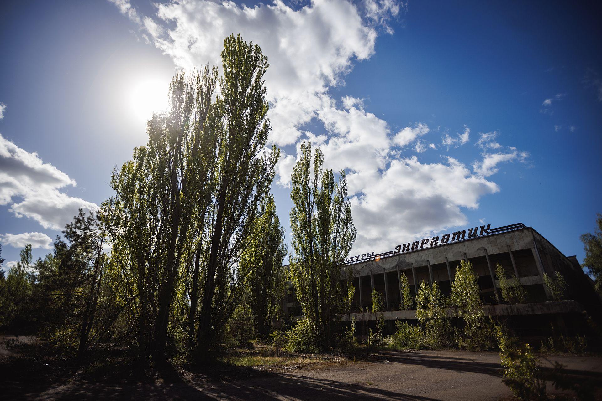 Vista exterior del abandonado Palacio de la Cultura Enerhetyk en la plaza central de Pripyat, óblast de Kiev, Ucrania, el 1 de mayo de 2025. Grandes árboles verdes crecen frente a él y se ve contra un cielo azul con nubes blancas.