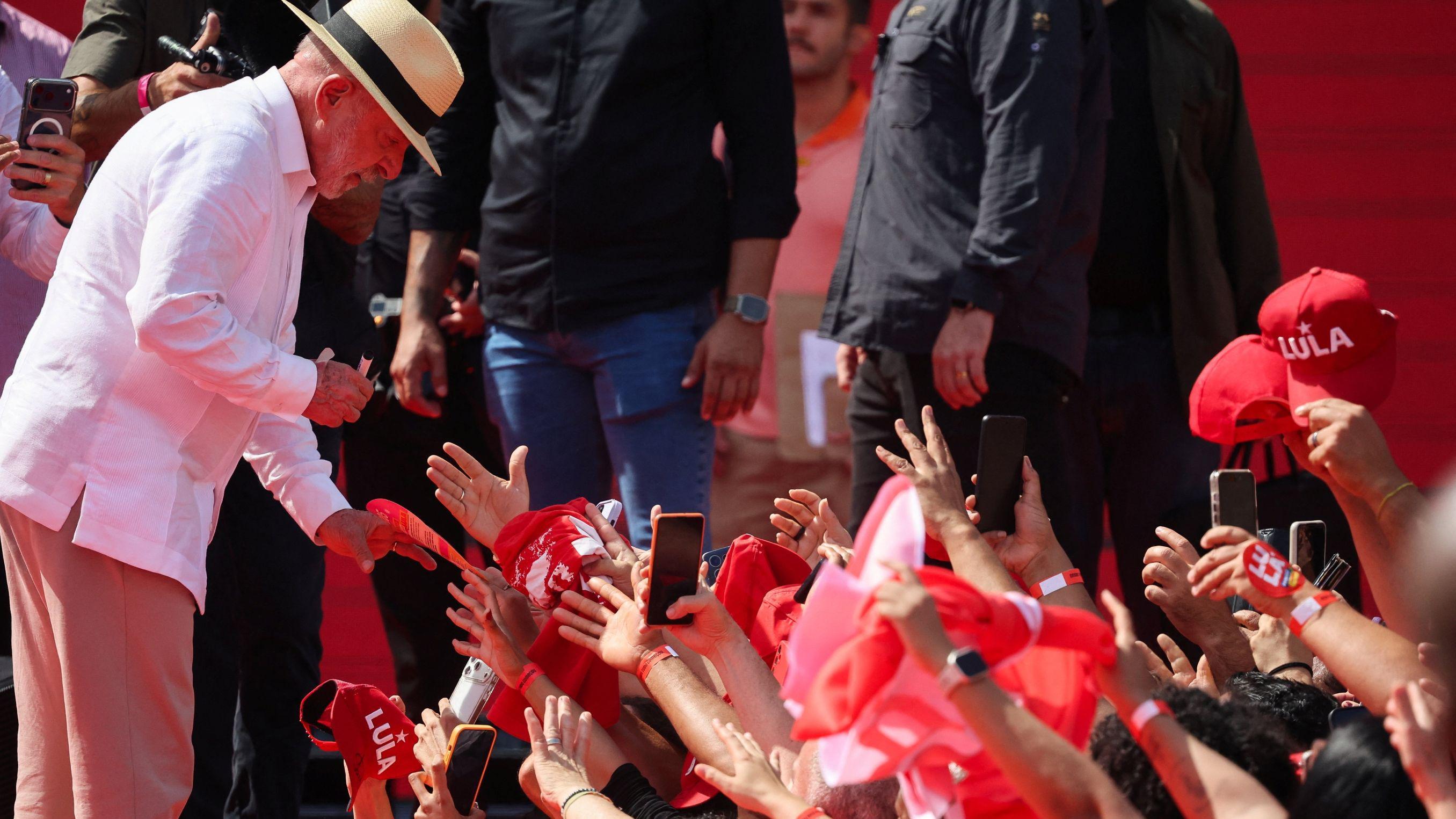 A imagem mostra Luiz In&aacute;cio Lula da Silva, de camisa e cal&ccedil;a brancas e usando chap&eacute;u, inclinado para cumprimentar apoiadores em um evento. Na plateia, v&aacute;rias pessoas estendem as m&atilde;os e seguram celulares, bandeiras e bon&eacute;s vermelhos com refer&ecirc;ncias a Lula.