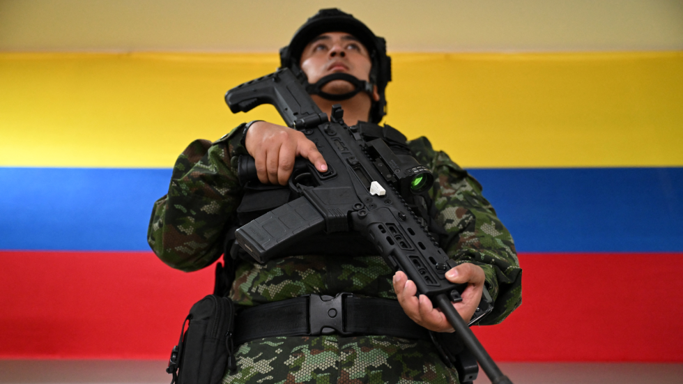 Un soldado sostiene un arma. Esta de pie delante de una bandera de Colombia roja, amarilla y azul.
