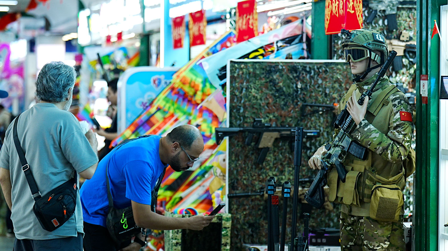 兩名男子看著一個攤位，攤位上有一名真人大小的玩具士兵拿著玩具槍站崗。