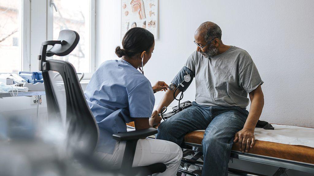 Profissional de assist&ecirc;ncia m&eacute;dica mede a press&atilde;o arterial de paciente sentado em uma maca hospitalar