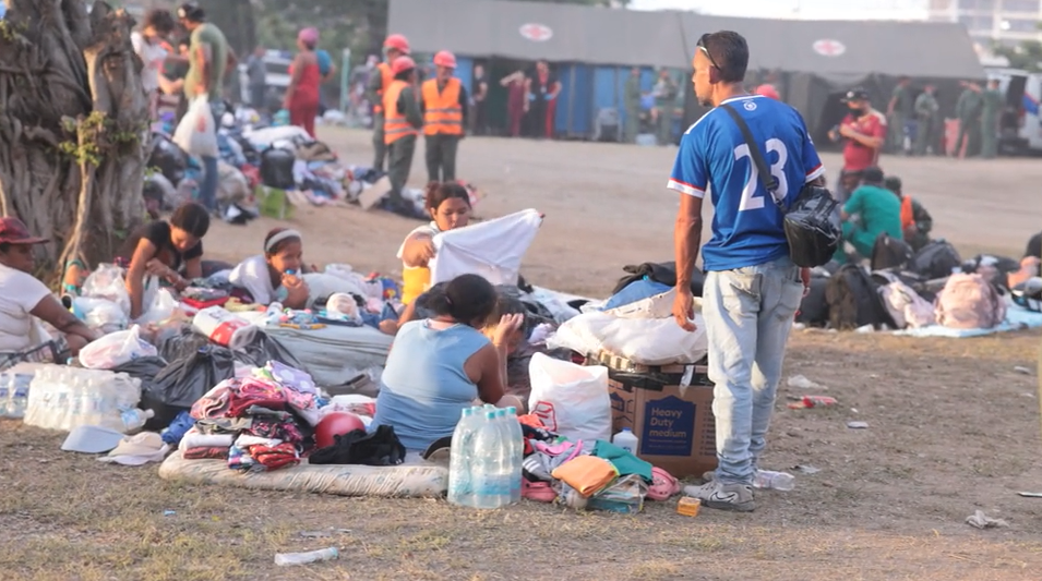 As pessoas se sentam no ch&atilde;o, cercadas por seus pertences, com uma tenda de emerg&ecirc;ncia ao fundo.