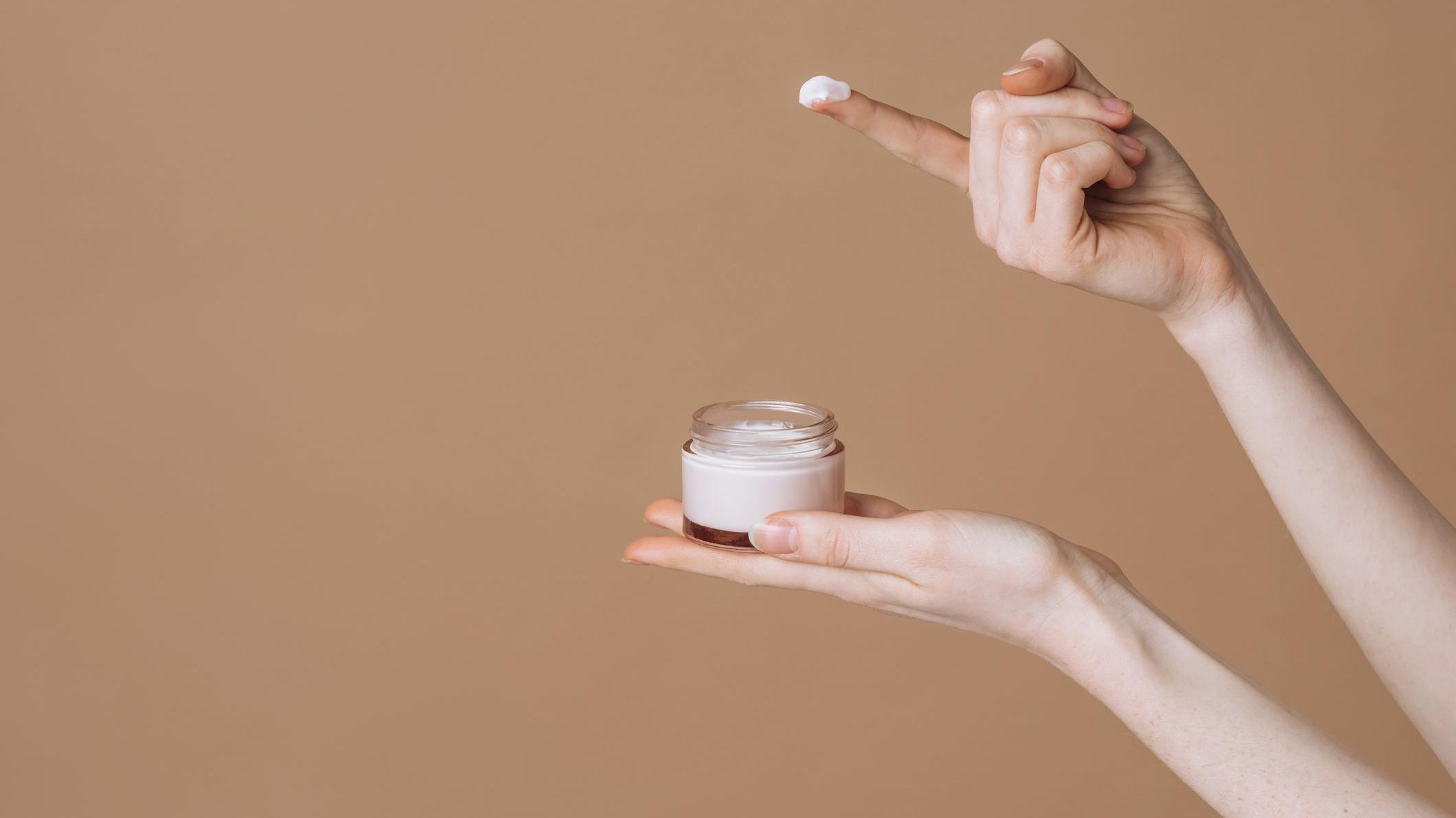 Uma m&atilde;o segura um pote de creme, enquanto o conte&uacute;do &eacute; retirado com o dedo de outra m&atilde;o.